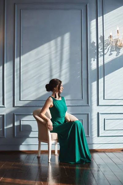Модная и красивая брюнетка-модель со стильной прической в зеленом вечернем платье сидит в кресле в роскошном интерьере в летний солнечный день — стоковое фото