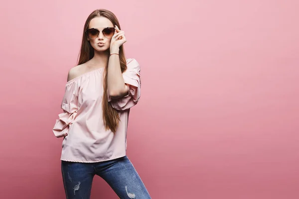 Menina loira modelo bonita e sexy em uma blusa com ombros nus e óculos de sol rosa na moda enviando beijo de ar e posando em um fundo rosa — Fotografia de Stock