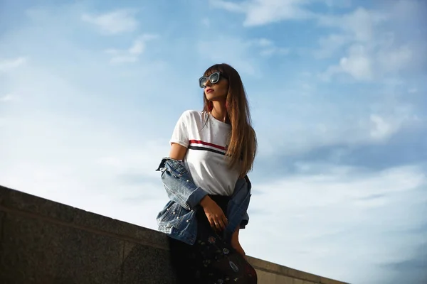Красивая и молодая модель женщина с идеальным телом в джинсовой куртке и в модной футболке позирует с голубым летним небом на заднем плане — стоковое фото