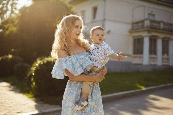 Hermosa y sorprendida joven rubia en vestido azul y amarillo de moda con un pequeño bebé lindo en sus manos —  Fotos de Stock