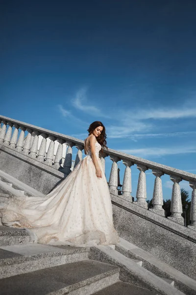 Menina morena bonita, sexy e elegante em vestido de noiva de renda branca posando nas escadas com céu azul de verão no fundo — Fotografia de Stock