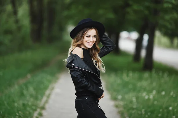 Mujer modelo joven de moda y sexy en una chaqueta de cuero negro y en jeans de moda ajustando su elegante sombrero negro y posando al aire libre —  Fotos de Stock