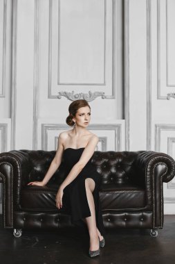 Şık ve zarif esmer model kız nazik makyaj büyük küpe ile, uzun siyah elbise, vintage kanepe üzerinde oturur ve lüks iç poz