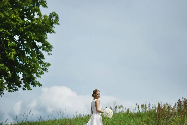 Bella giovane donna bionda in un abito bianco elegante con acconciatura di nozze mantenendo un mazzo di fiori bianchi e in posa all'aperto in un campo verde. Giovane sposa che cammina prima della cerimonia nuziale — Foto Stock