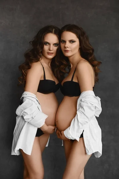 Two beautiful pregnant twins model girls in sexy black lingerie and oversized shirts. Two young gestation women with perfect bodies stay close to each other, posing at the dark grey background — Stock Photo, Image