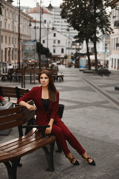 Fashionable and young pretty woman, in a cherry smart suit, sitting on the bench and relaxing outdoors. Stylish brunette model girl posing on a square and looking aside. Concept of city life and style
