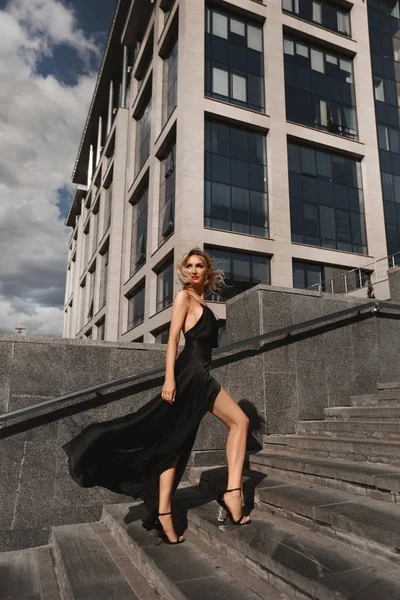 Beautiful, sexy and fashionable blonde girl with perfect body in black evening dress posing at urban background — Stock Photo, Image