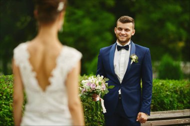 Şık ve sakallı genç vahşi adam akıllı bir takım elbise buket tutan ve bir gelinlik mükemmel bir vücut ile onun seksi ve güzel model kız bakıyor, yeşil parkta açık havada