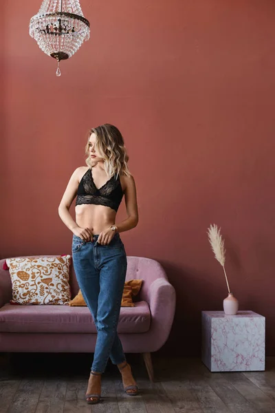 Schöne und sexy blonde junge Frau, mit einem schlanken perfekten Körper, in Jeans und modischem schwarzen Spitzen-BH, der sich auszieht und im Wohnzimmer posiert — Stockfoto