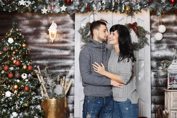 Bella coppia di amanti, elegante bell'uomo che abbraccia con bella bruna giovane donna in una felpa grigia alla moda e posa vicino all'albero di Natale all'interno decorato per il nuovo anno — Foto Stock