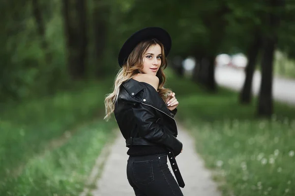 Ragazza modello bionda emotiva e alla moda con trucco luminoso, in una giacca di pelle nera, cappello alla moda e jeans in posa all'aperto per la strada della città. Concetto di street fashion . — Foto Stock