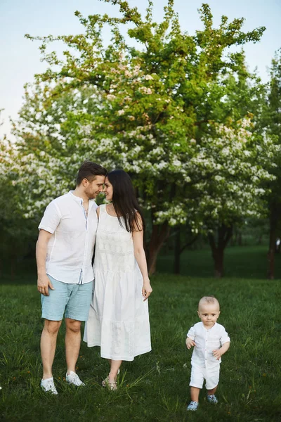Bei giovani genitori, elegante bell'uomo in pantaloncini e camicia e bellezza bruna nel vestito bianco, e il loro piccolo figlio carino si divertono vicino agli alberi al parco in fiore. Concetto di famiglia felice — Foto Stock