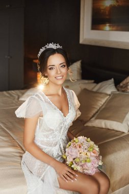 Beyaz iç çamaşırı ve modish peignoir güzel seksi genç esmer gelin yatak odasında yatakta oturur. Mükemmel vücut ve parlak gülümseme ile model kız iç düğün buketi ile poz