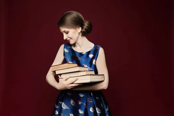 Estudante feminina trabalhadora em vestido azul, mantendo os livros nas mãos e olhando para a câmera. Menina bonita com olhos azuis e penteado na moda posando com uma pilha de livros em fundo vermelho, isolado — Fotografia de Stock