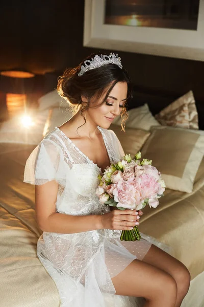 Bella giovane sposa bruna sexy in lingerie bianca e peignoir modish si siede sul letto in camera da letto. Ragazza modello con il corpo perfetto, in abito sexy, in posa con bouquet da sposa in interni — Foto Stock