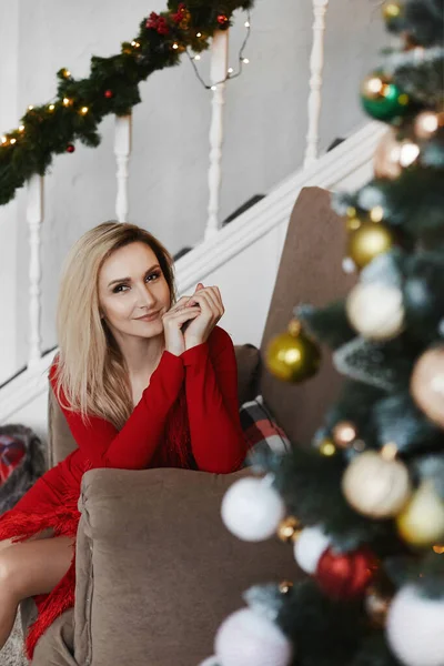 Mujer joven rubia en vestido rojo modesto posando cerca del árbol de Navidad en el interior decorado para Año Nuevo —  Fotos de Stock