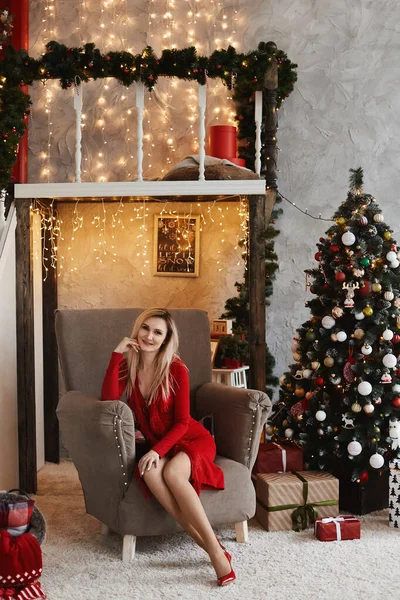 Mujer joven rubia en vestido rojo modesto posando cerca del árbol de Navidad en el interior decorado para Año Nuevo —  Fotos de Stock