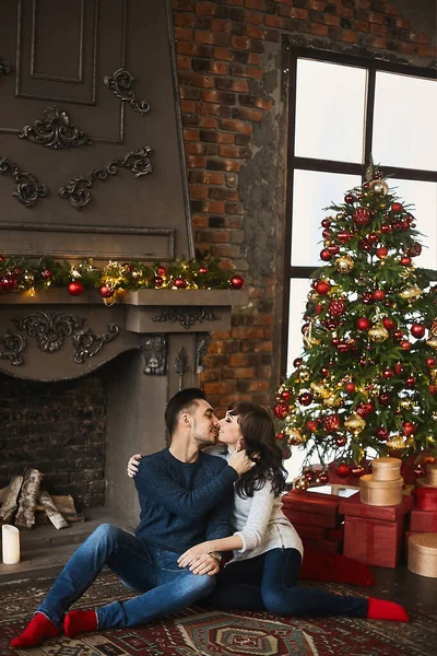 Le jeune couple heureux, en chandails chauds et chaussettes rouges, étreignant et assis sur le tapis authentique près de la cheminée et de l'arbre de Noël. Célébration de Noël et Nouvel An — Photo