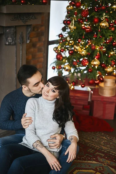Le jeune couple heureux, en pull chaud. Jeune bel homme embrassant une belle petite amie près de la cheminée et arbre de Noël en arrière-plan. Célébration de Noël et Nouvel An — Photo