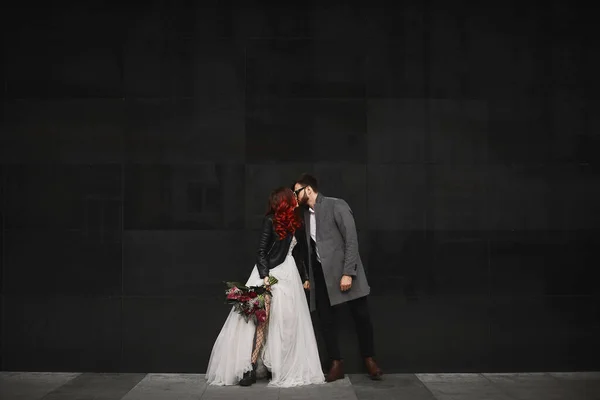 Beautiful couple of lovers kisses outdoors during wedding photoshoot. Slim young woman with red hair in modish leather jacket and wedding dress and handsome bearded man in coat. — Stock Photo, Image