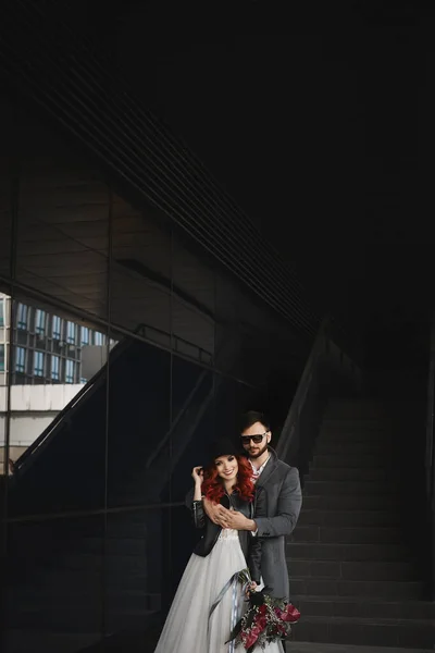 Beautiful couple of lovers hugging outdoors during wedding photoshoot. Slim young woman with red hair in modish leather jacket and wedding dress and handsome bearded man in the coat. — Stock Photo, Image