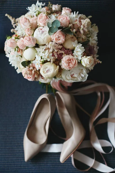 Komposition aus dem Brautstrauß wunderschöner Pfingstrosen-Blumen mit einem Paar modischer Brautschuhe auf dunkelblauem Hintergrund. Konzept der Hochzeitsvorbereitung. Accessoires für eine Braut — Stockfoto