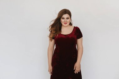 Beautiful plus size model girl in modish red velvet dress isolated at white background. Young fat woman with bright makeup and with stylish hairstyle posing in studio. Concept of XXXL fashion. clipart
