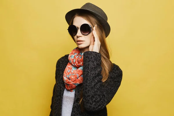 Una hermosa joven con abrigo modesto, sombrero de moda y gafas de sol posando en el fondo amarillo, aislada. Modelo chica con el pelo rubio en traje de otoño modesto. Concepto de moda callejera —  Fotos de Stock