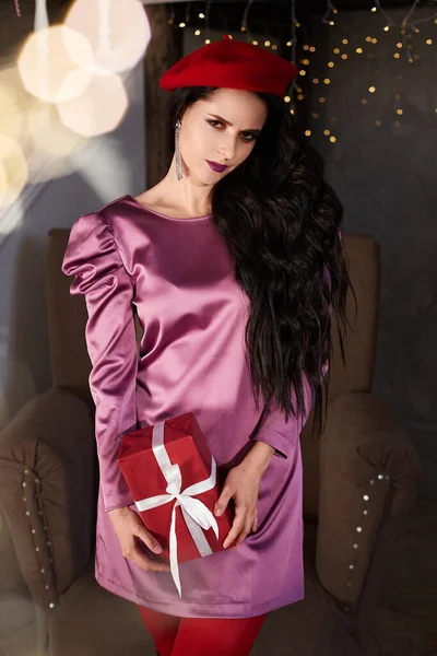 Sensuele en mooie jonge vrouw met heldere make-up, in modische roze jurk en rode baret, houden kerstcadeau en poseren in het interieur. Een brunette model meisje in modieuze outfit. — Stockfoto