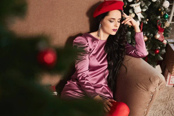 Sensuele en mooie jonge vrouw met heldere make-up en gesloten ogen, in modische roze jurk en rode baret, zit in de buurt van kerstboom. Een brunette model meisje in modieuze outfit. — Stockfoto