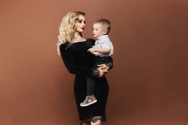 Siyah elbiseli, güzel ve mutlu genç sarışın kadın ellerinde küçük bir oğlan çocuğu, arka planda soyutlanmış. Modern annelik kavramı.