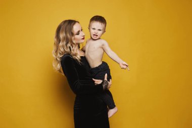 Siyah elbiseli, güzel ve mutlu sarışın bir kadın. Elinde küçük bir oğlan çocuğu var. Sarı arka planda izole edilmiş. Modern annelik kavramı.
