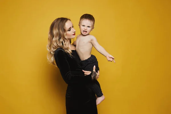 Beautiful and happy young blonde woman in a black dress with a cute little baby boy on her hands, isolated at the yellow background. Concept of modern maternity.