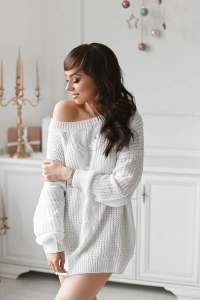 Mujer morena joven atractiva y de moda con maquillaje brillante y peinado elegante, en suéter blanco y medias posando en el interior decorado para Año Nuevo. Concepto de la mañana de Navidad —  Fotos de Stock