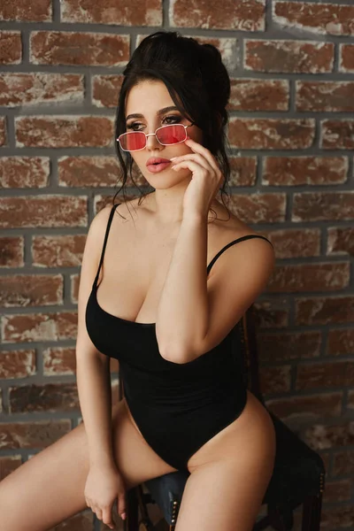 Ung brunett kvinna med en förförisk stor byst i body suits och glasögon med röda linser sitter och poserar nära tegelväggen. Sensuell bystig modell flicka med perfekt kropp i modisk outfit. — Stockfoto