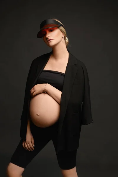 Высокомодная молодая беременная женщина в модном черном наряде с обнаженным желудком позирует и смотрит в камеру на тёмном фоне, изолированная. Модная беременная девушка-модель на последнем месяце беременности — стоковое фото