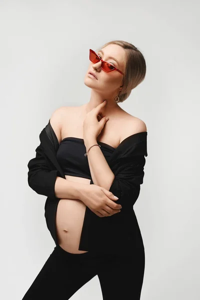 Высокомодная беременная модель девушка в модном черном наряде с обнаженным животом позирует и смотрит в камеру на белом фоне, изолированный. Модифицированная беременная молодая женщина на последнем месяце беременности — стоковое фото