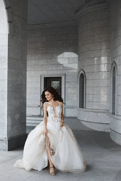 Een gelukkige jonge bruid met een trouwkapsel in een witte kanten jurk loopt door de binnenplaats van een oude kerk. mooi model meisje houdt haar bruiloft jurk in haar handen en poseert in de oude stad — Stockfoto