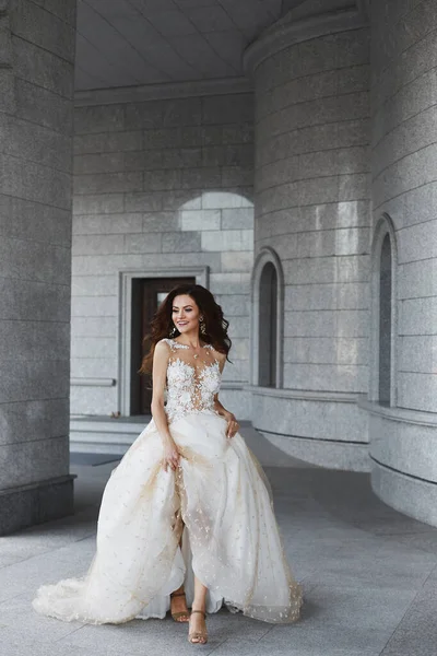Een gelukkige model vrouw met een bruiloft kapsel in een witte kant jurk loopt door de binnenplaats van een oude kerk. Mooie jonge bruid houdt haar trouwjurk in haar handen en poseert in de oude stad — Stockfoto
