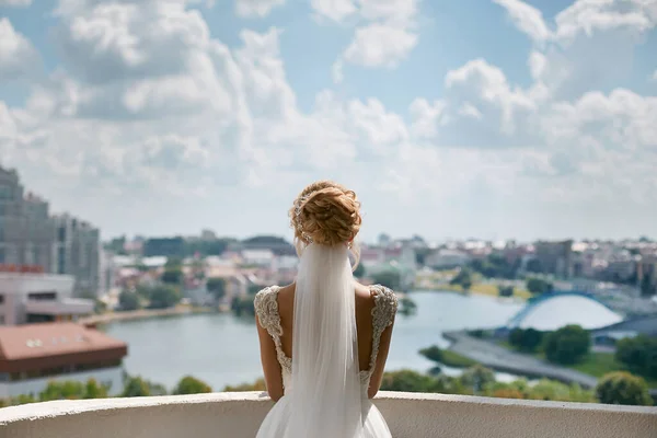 Elegante mujer joven con peinado elegante, en vestido de novia de encaje y velo nupcial mira a la vista de la ciudad, la preparación de la boda de la novia joven — Foto de Stock