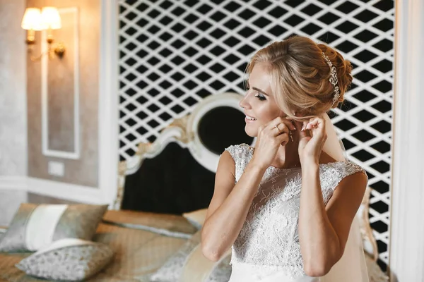 Modische und elegante blonde Model-Mädchen mit stilvoller Hochzeitsfrisur, im Spitzenkleid, setzt ihren Ohrring auf und posiert im Inneren — Stockfoto