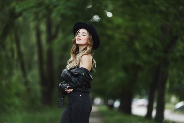 Deri ceketli ve siyah şapkalı güzel bir kızın yeşil bir caddede yaz gününün tadını çıkarırken görüntüsü..