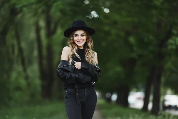 一个穿着皮夹克、头戴黑帽的年轻女子独自走在夏季城市街道上的画像 — 图库照片