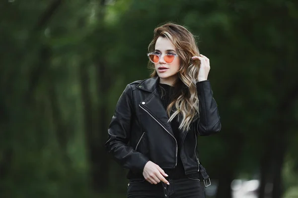 Young beautiful girl in a leather jacket and fashionable sunglasses is walking alone outdoors in summer day — Stock Photo, Image