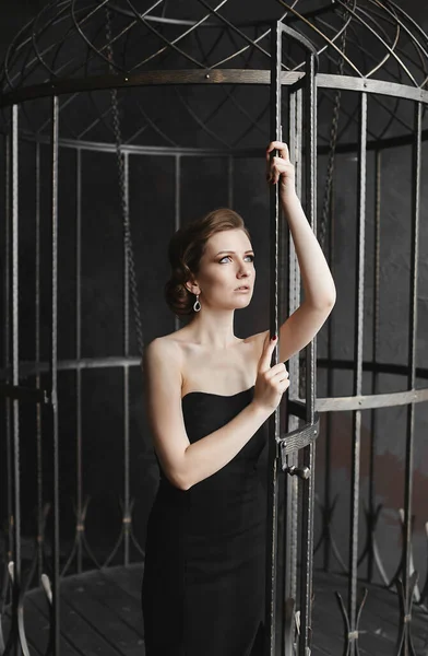Μια νεαρή όμορφη γυναίκα με μαύρο φόρεμα που ποζάρει κοντά στο κλουβί. Σύμβολο της ελευθερίας και το τέλος της απομόνωσης. — Φωτογραφία Αρχείου
