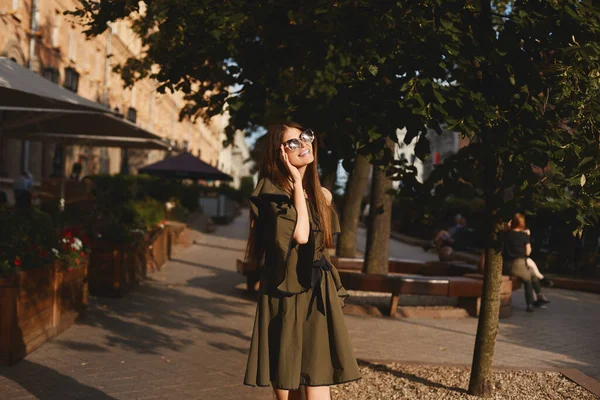 Portrait of a cheerful young woman in dress touching her sunglasses on urban background. Fashionable girl with beautiful brown hair smiling to camera. — Stock Photo, Image