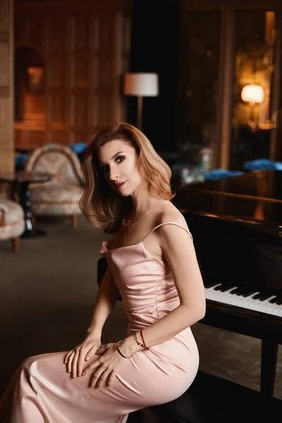 Bej bir gece elbisesi içinde mükemmel vücutlu manken kız lüks bir iç mekanda büyük piyanonun başında oturuyor.. — Stok fotoğraf
