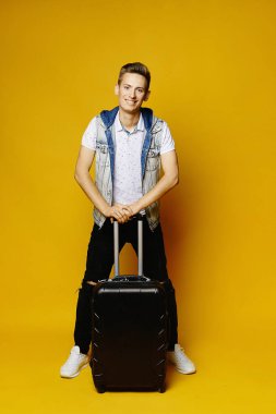 Genç yakışıklı bir adam bir seyahat çantası tutuyor ve tatile hazır, sarı bir arka planda izole edilmiş..