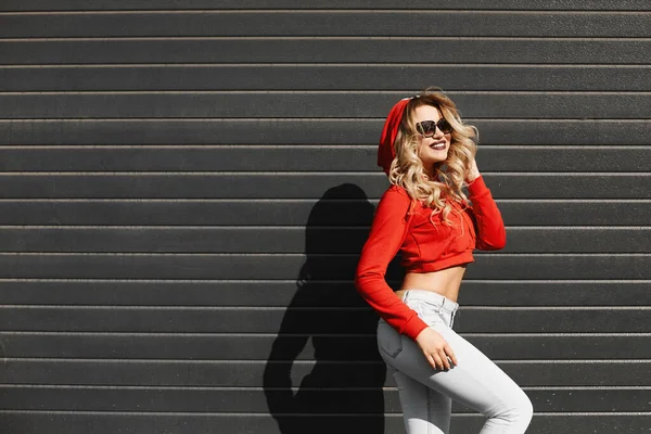 Model girl wearing a red hoodie, jeans and hipster sunglasses posing against street wall, urban clothing style. — Stock Photo, Image