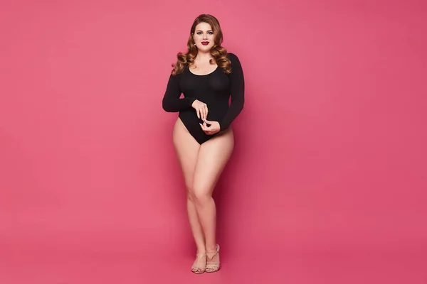 Jovem mulher gorda com maquiagem e penteado na moda vestindo um bodysuit preto e posando no fundo rosa, isolado . — Fotografia de Stock
